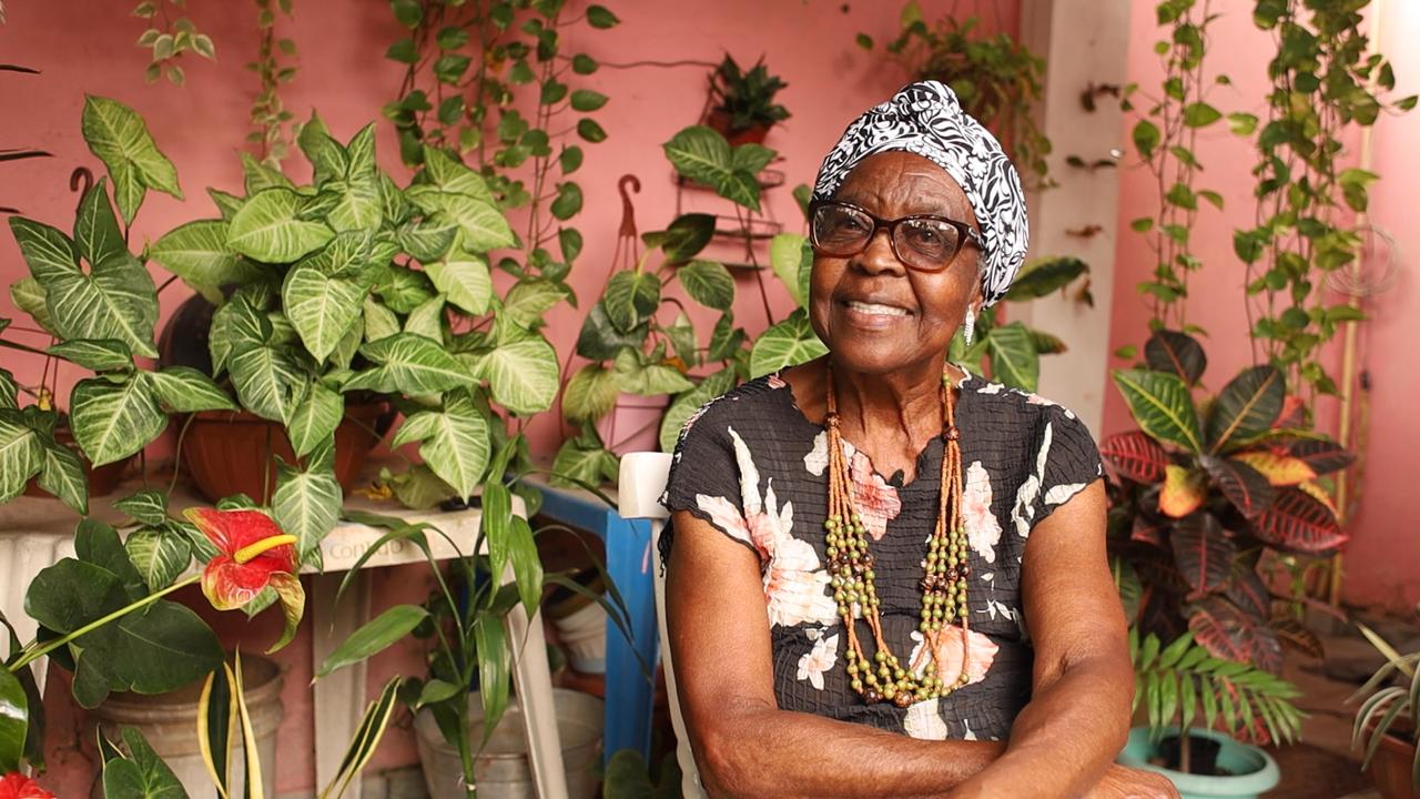 Dona Zica, uma das entrevistadas do filme, sorri para foto em um quintal cheio de plantas. Dona Zica é uma mulher negra, usa óculos, turbante preto e branco nos cabelos, um vestido florido e colar de contas.