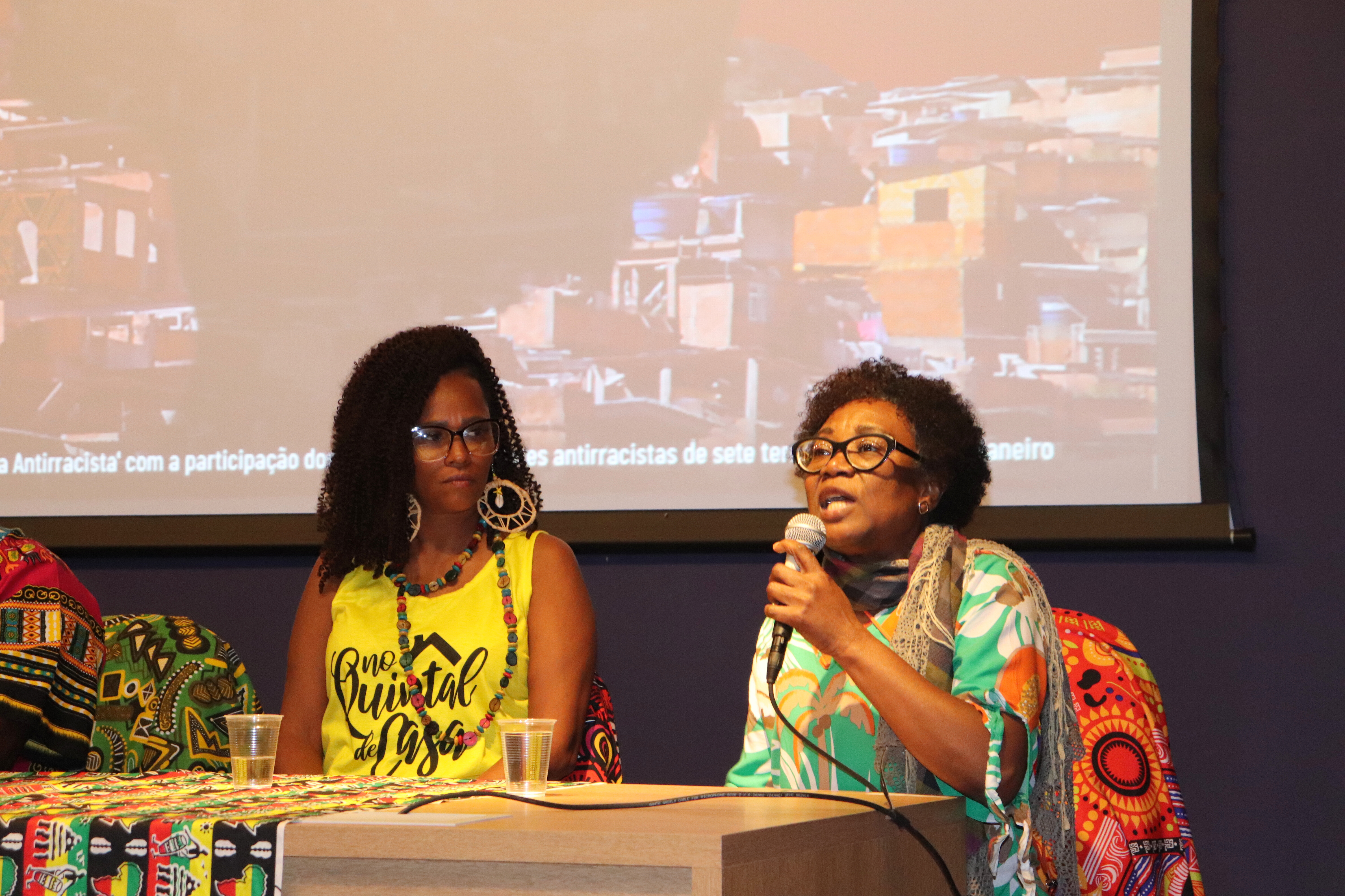 Cláudia Lima realiza sua fala durante o encerramento da segunda mesa de debate. Cláudia é uma mulher negra de cabelos crespos curtos, usa poculos de grau e veste uma encharpe cinza por cima de uma blusa verde estampada.