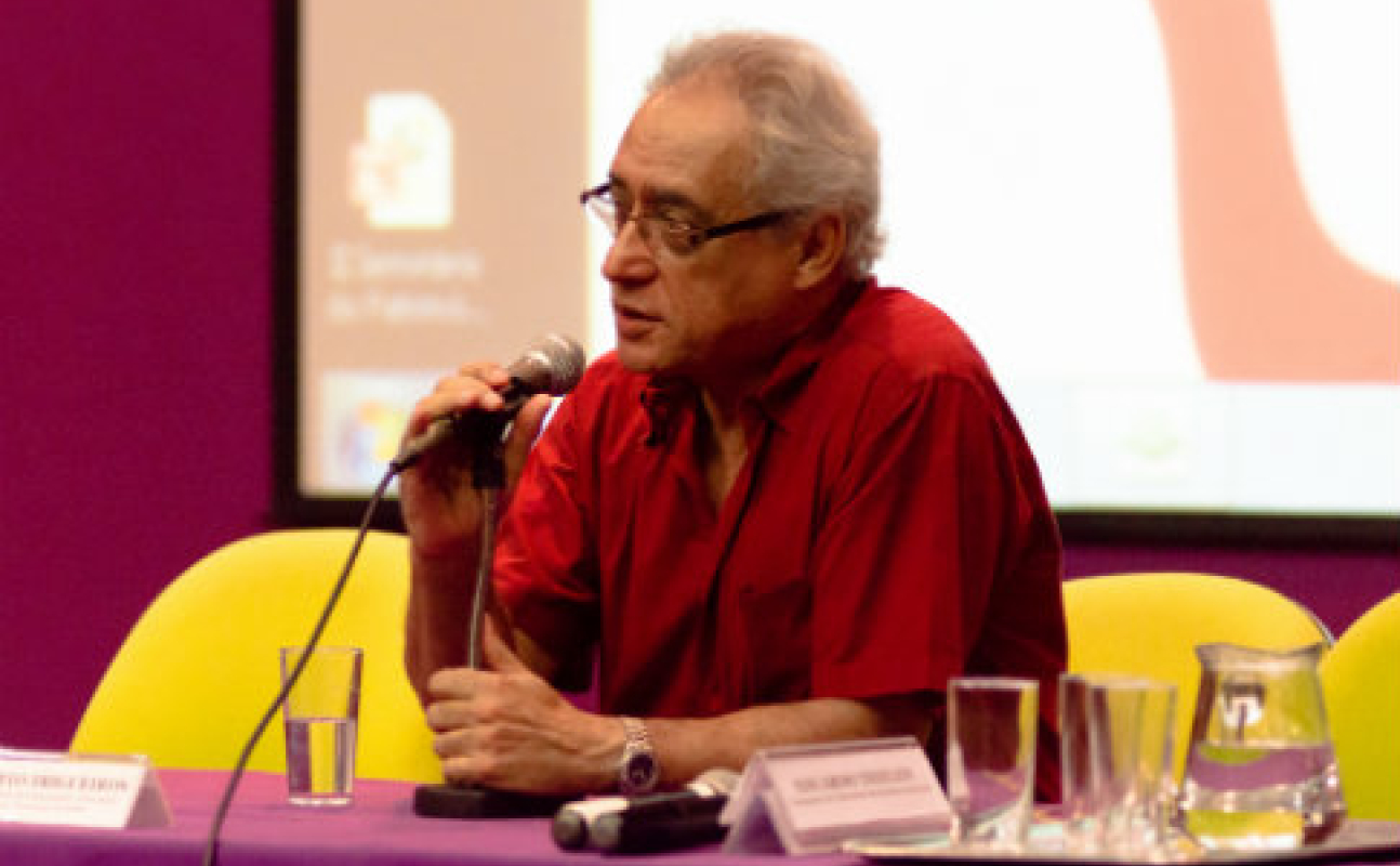 Foto do diretor em mesa, falando ao microfone com projeção ao fundo