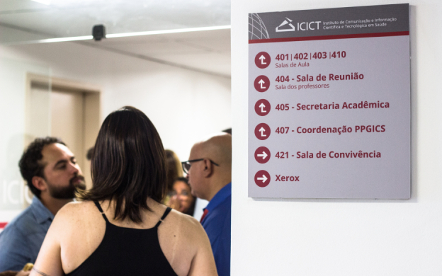 Foto de acadêmicos conversando com destaque para placa de indicações nos corredores do PPGICS. Foto: Raquel Portugal