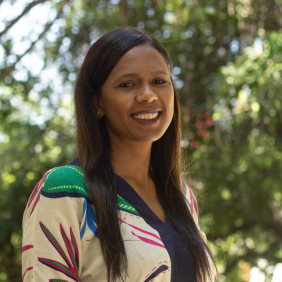 Pesquisadora Viviane Santos de Oliveira Veiga, mulher, negra, cabelos longos, camisa estampada, em fundo ambiente com vegetação. Foto: Raquel Portugal/Multimeios.