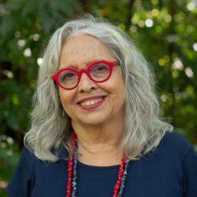 Pesquisadora Inesita Araujo, parda, cabelos grisalhos, óculos vermelho, camisa azul, com fundo de natureza. Foto: Raquel Portugal/Fiocruz Imagens. 