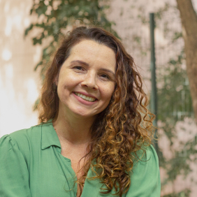 Pesquisadora Izamara Bastos, branca, cabelos cacheados longos louros, camisa verde,  com fundo de natureza. Foto: Raquel Portugal/Fiocruz Imagens. 