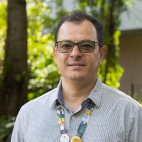 Pesquisador Marcel Pedroso, homem branco, cabelos pretos curtos, óculos preto, camisa quadriculada azul, com árvores ao fundo. Foto: Raquel Portugalcamisa 