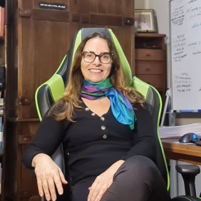Pesquisadora Cristina Rabelais, branca, cabelos louros compridos, branca, de óculos, camisa preta, lenço no pescoço, sentada em frente à escrivaninha, com armários ao fundo. Foto: divulgação Nippis