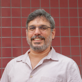 Pesquisador Miguel Papi, branco, camisa social, cabelo e barba grisalhos, de óculos, com fundo de ladrilho vermelho