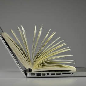 Livro com páginas abertas, apoiado como se conteúdo tivesse saindo de um notebook. 