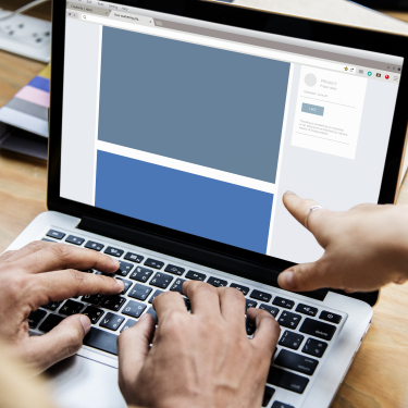 Pessoa aponta para a tela de um notebook enquanto outra usa o teclado, visualizando o layout de uma página web em desenvolvimento.