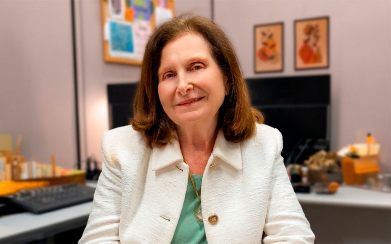 Foto da pesquisadora Celia Szwarcwald, mulher, branca, cabelos lisos na altura do pescoço, com camisa verde e casaco branco, em sala de trabalho