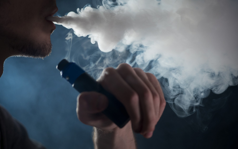 Pessoa utilizando cigarro eletrônico e exalando grande nuvem de vapor em ambiente escuro. Imagem: Freepik.