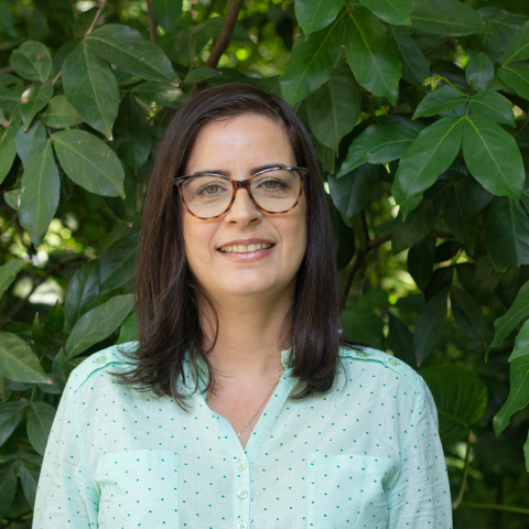 Pesquisadora Giseli Damacena, branca, mulher, cabelos castanhos lisos na altura do ombro, óculos e camisa verde.