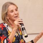 Pesquisadora branca, cabelo loiro, com camisa estampada, segurando o microfone.