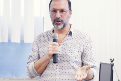 Pesquisador Josue Laguardia, branco, de barba, óculos, camisa branca com pontinhos, com microfone na mão. Foto: PPGICS.