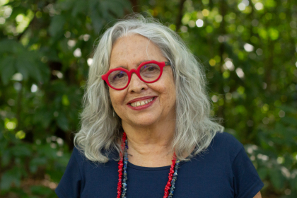 Pesquisadora Inesita Araujo, parda, cabelos grisalhos, óculos vermelho, camisa azul, com fundo de natureza. Foto: Raquel Portugal/Fiocruz Imagens. 