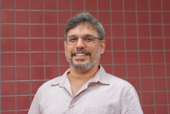 Pesquisador Miguel Papi, branco, camisa social, cabelo e barba grisalhos, de óculos, com fundo de ladrilho vermelho